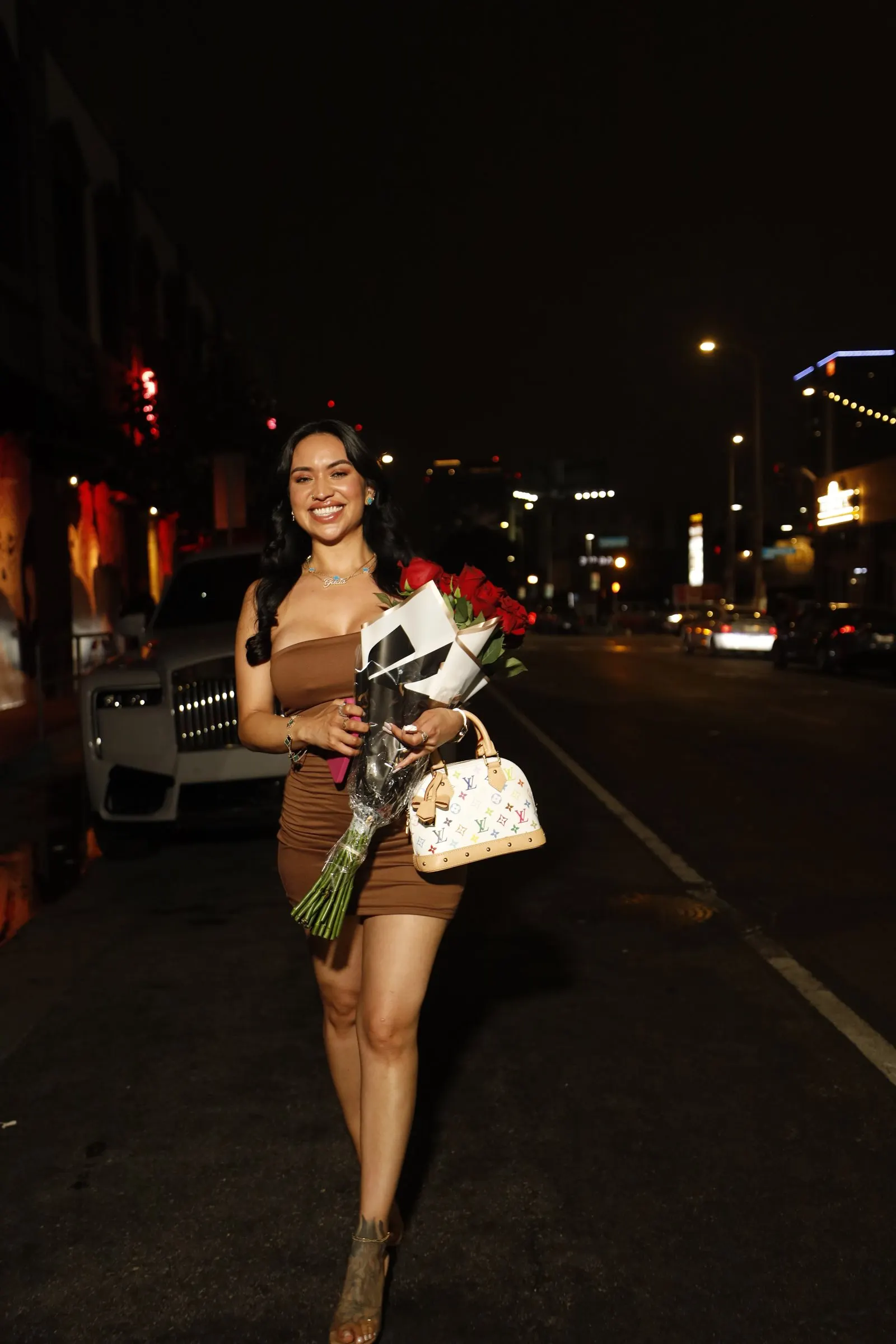 A guest in a brown mini dress arrives at Koza Hollywood carrying red roses and a monogram handbag